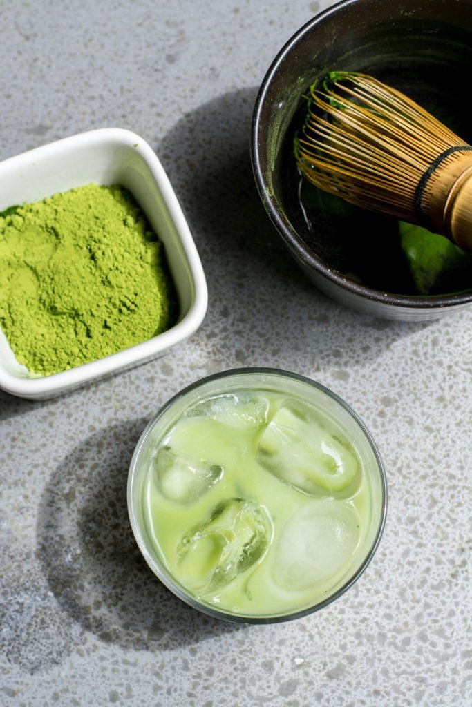 Matcha powder, a black chawan with bamboo whisk, and an iced matcha on a grey surface